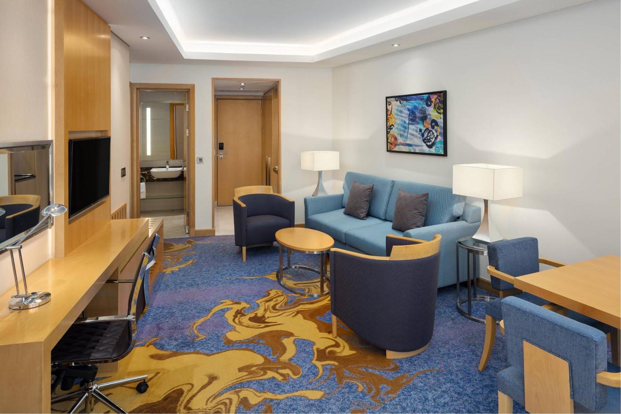 Sheraton Makkah Jabal Al Kaaba Hotel Mekka Exteriér fotografie The photo displays a modern hotel room or suite. The room features a comfortable seating area with a blue couch and two blue armchairs surrounding a small round table. A wooden desk with a black office chair is positioned against one wall, accompanie