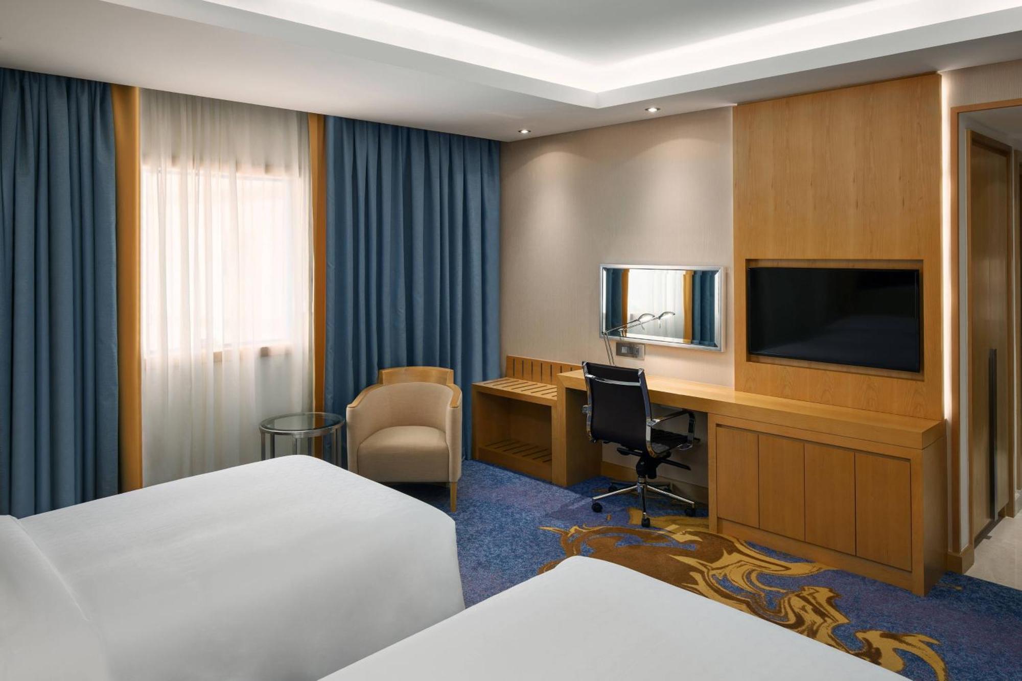 Sheraton Makkah Jabal Al Kaaba Hotel Mekka Exteriér fotografie The photo shows a hotel room featuring two beds with white linens. There are large windows covered with blue curtains that allow natural light to enter. On one side of the room, there is a wooden workstation with a chair and a mirror, along with a fl