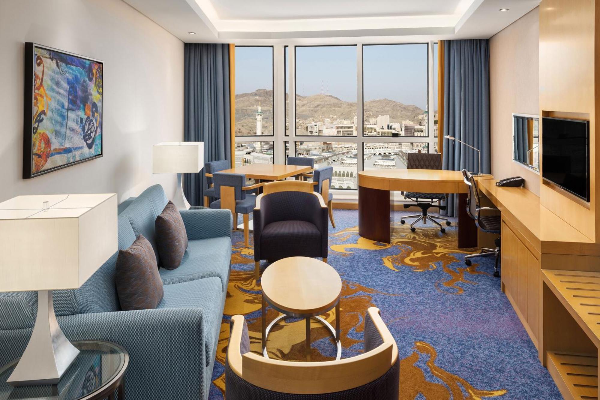 Sheraton Makkah Jabal Al Kaaba Hotel Mekka Exteriér fotografie The photo shows a well-decorated hotel room featuring a modern and comfortable interior. There is a cozy seating area with a blue sofa and armchairs, complemented by a round coffee table. A desk with a chair is also present, ideal for work. The room 